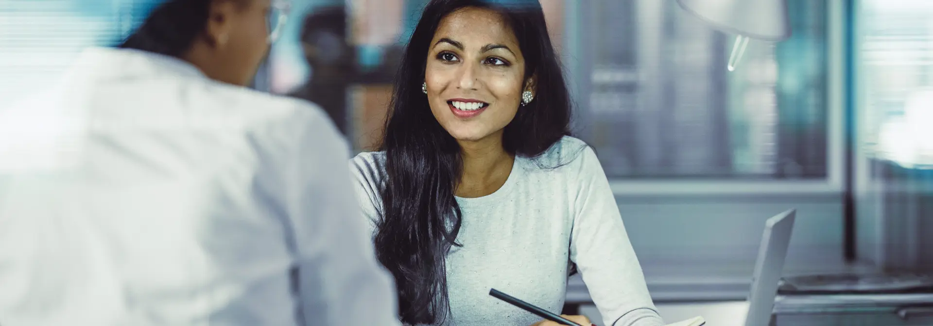 A woman sitting in an office smiles as she takes notes while in conversation.
