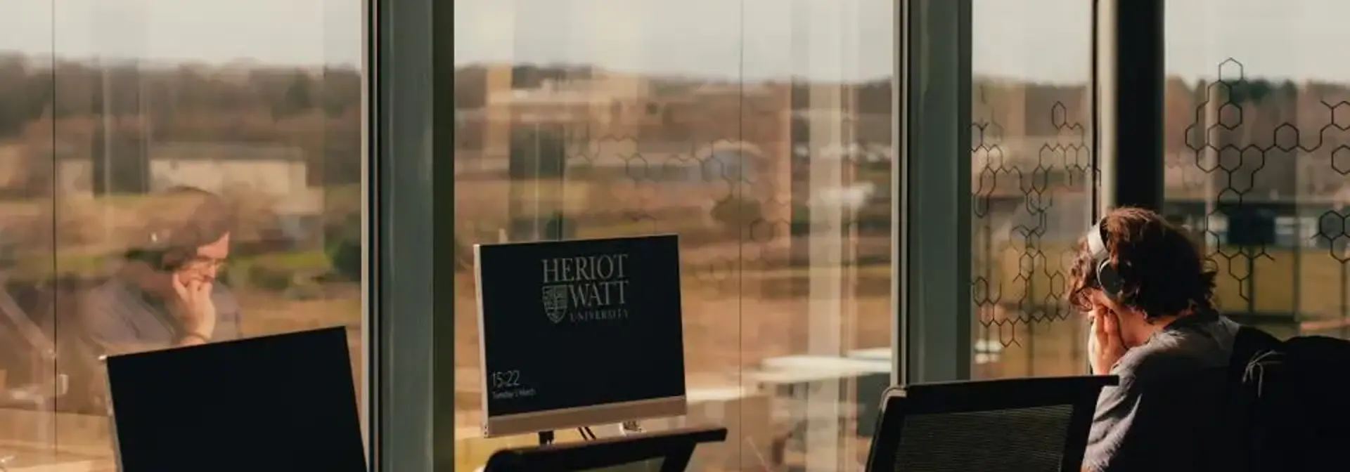 A man seated at a desk, working on a computer, with a large window providing natural light.