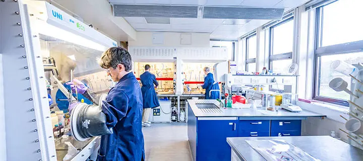 Two students in a chemistry lab, one using an isolation glove box and one talking with a member of staff