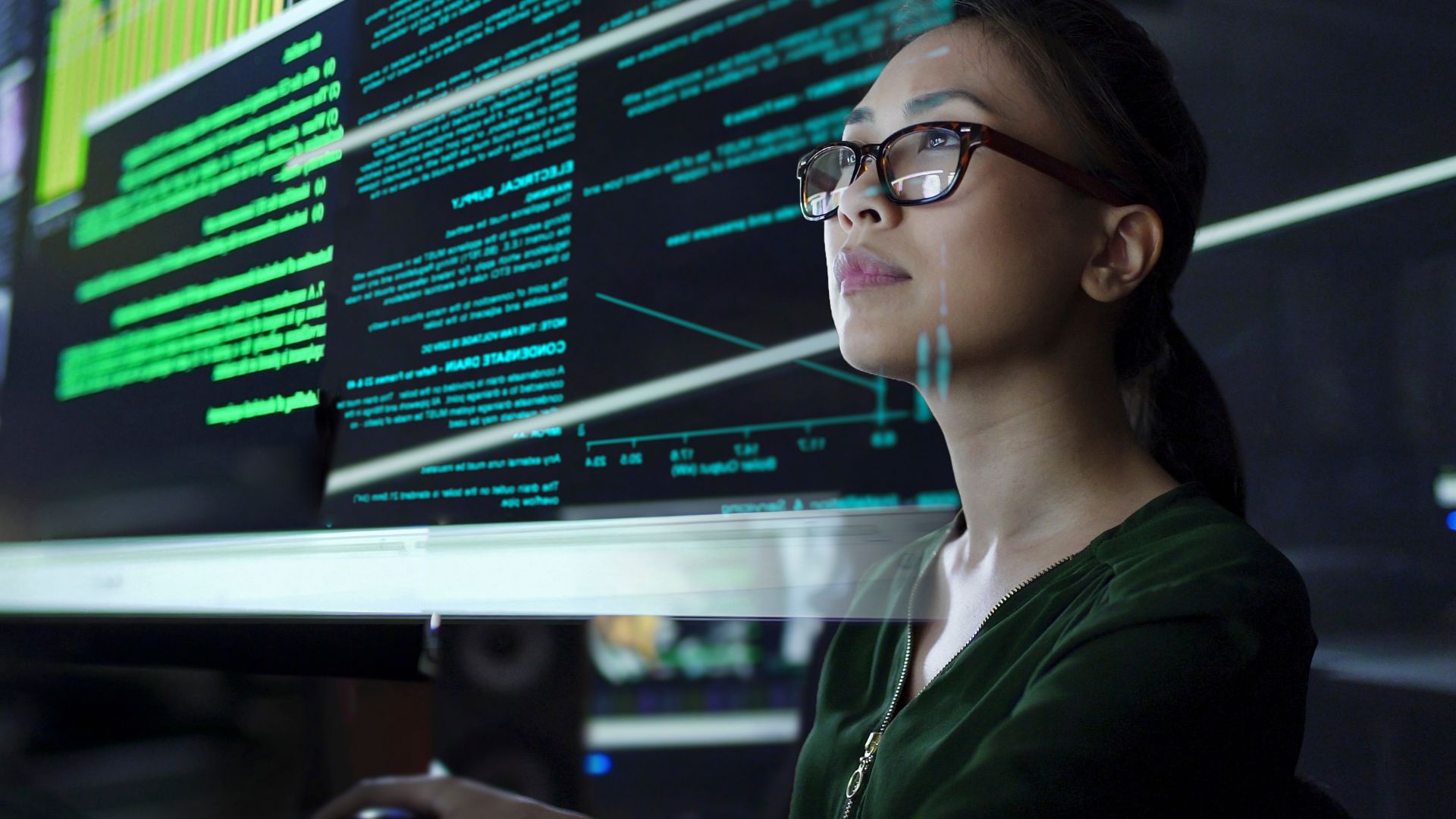 A woman wearing glasses studies computer code on a large screen