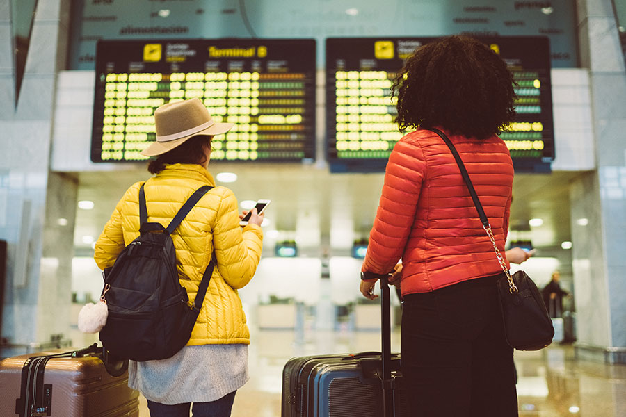 Two women with suitcases study an airport flight departure board