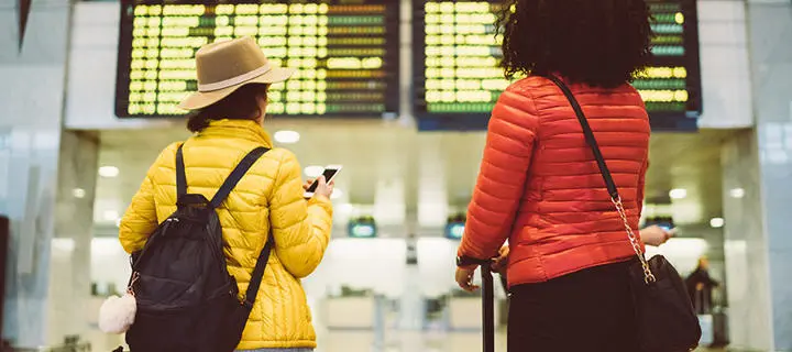 Two women with suitcases study an airport flight departure board