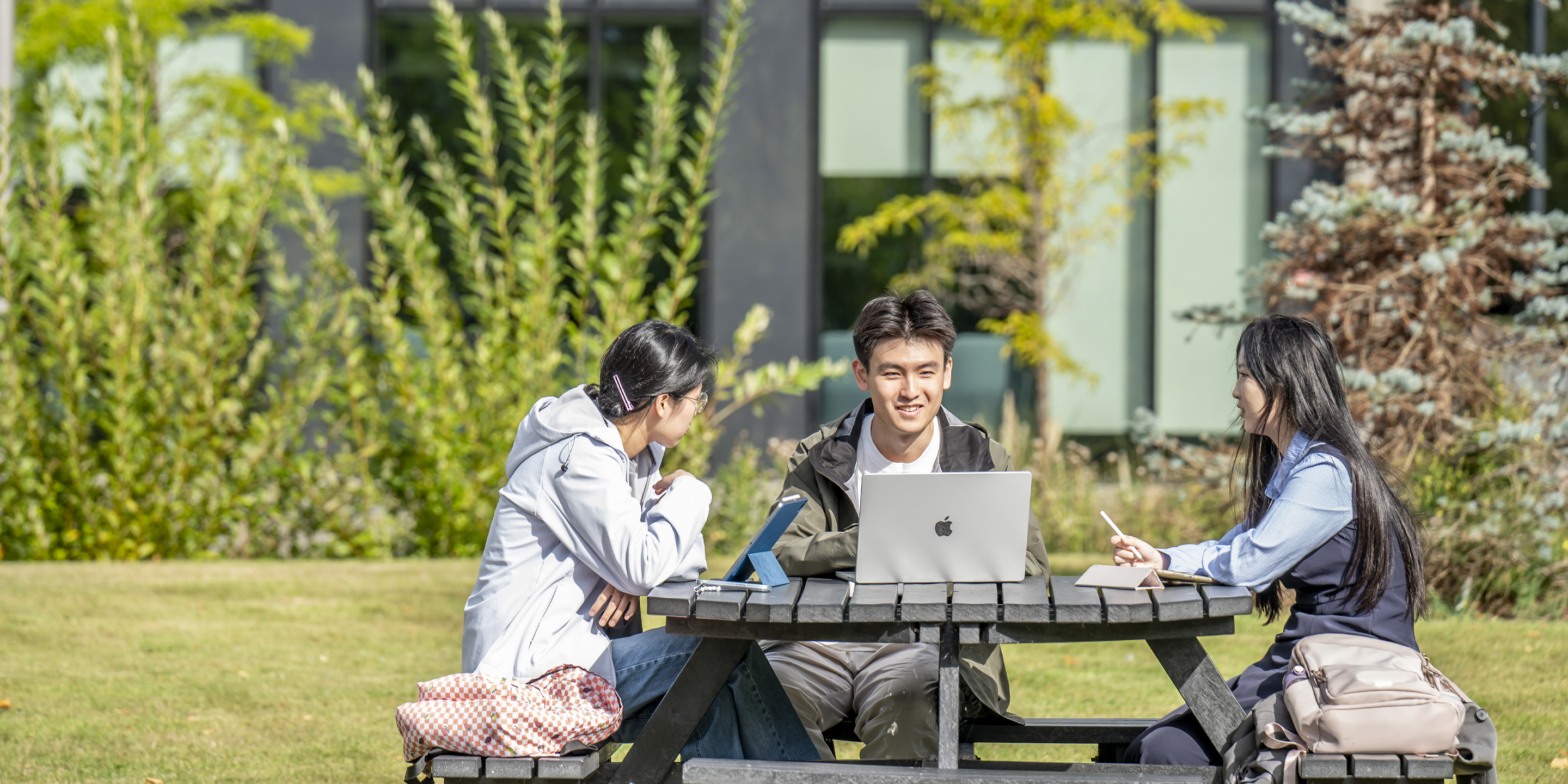Three students sitting at a picnic table outside, studying together.