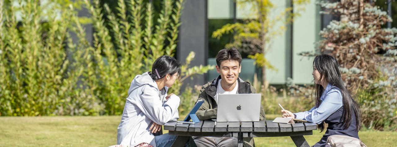 Three students sitting at a picnic table outside, studying together.