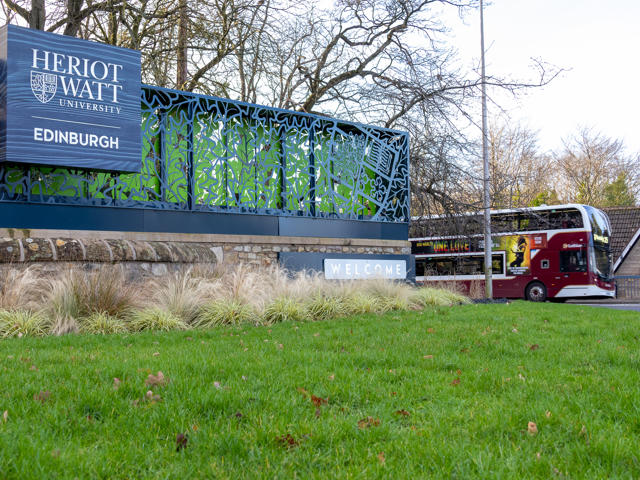 An entrance sign for Heriot-Watt University with a bus driving in the background.