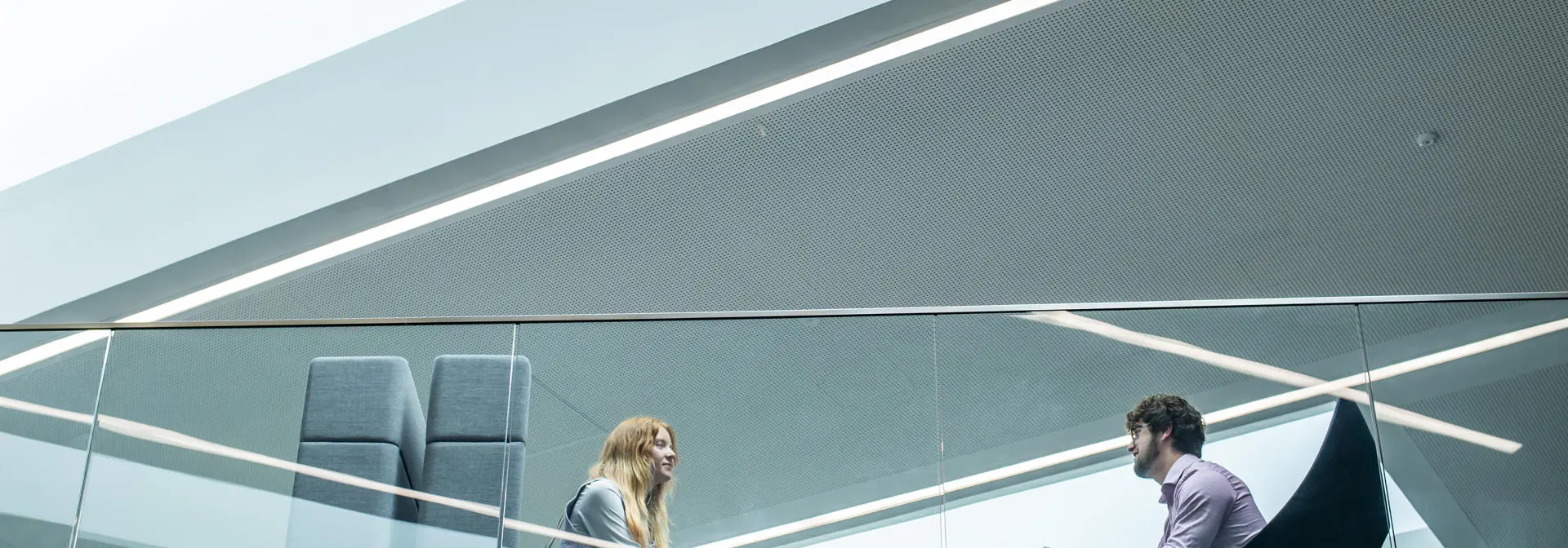 Two individuals sit opposite each other at a table and chairs, behind a glass barrier.
