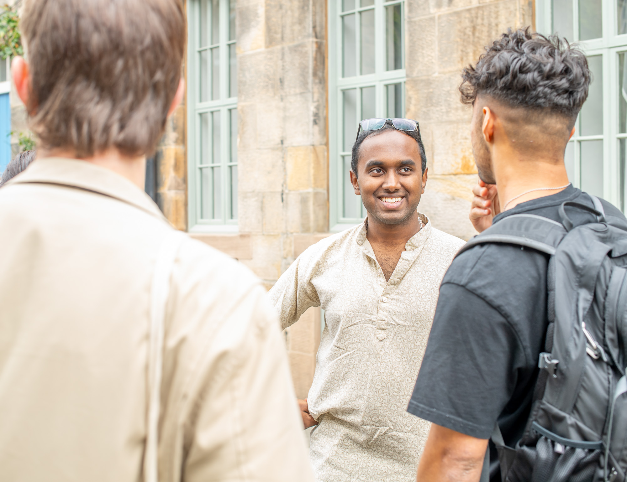 A smiling man speak with two individuals beside a stone wall.
