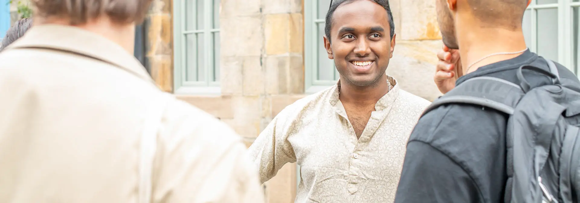 A smiling man speak with two individuals beside a stone wall.