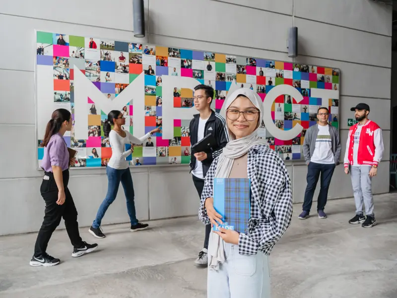 A woman stands in front of a colourful mural, people walking behind her as she smiles holding a book.