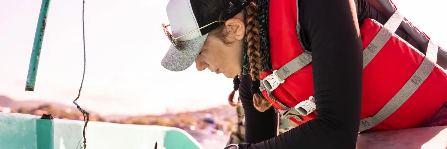 A student wearing a life jacket is on a boat writing notes.