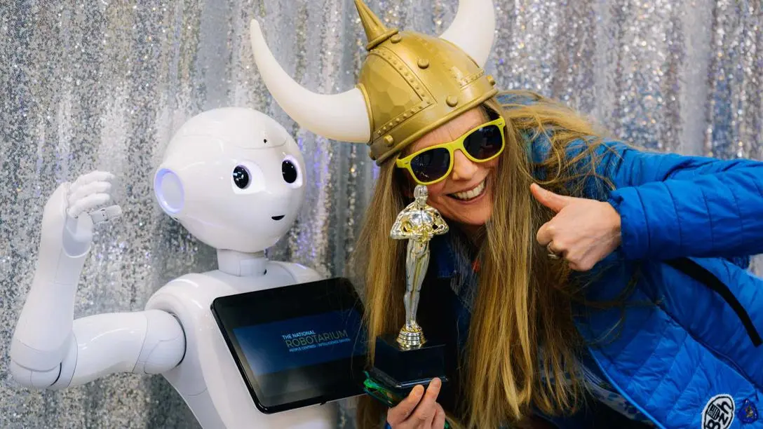 A blonde woman wearing a Viking hat and sunglasses stands smiling beside a humanoid robot