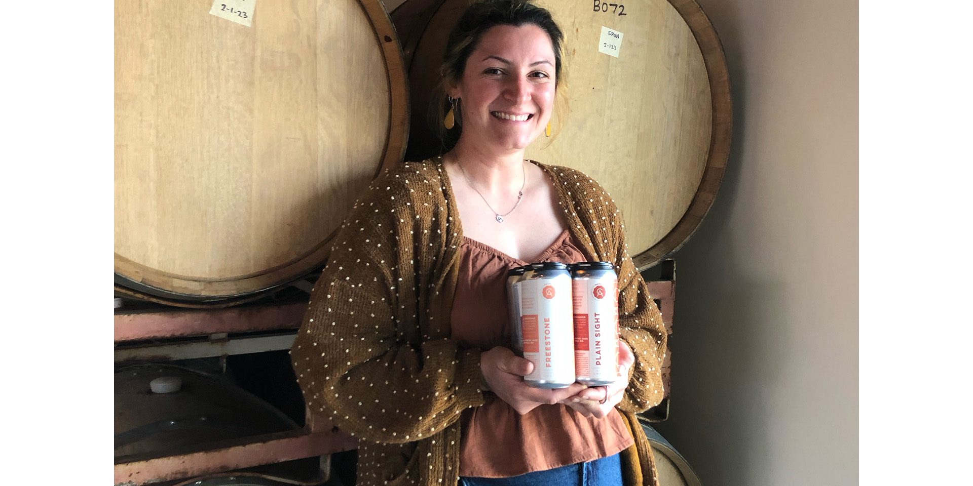 Bella Perez standing in front of two beer barrels, smiling, holding two cans of beer