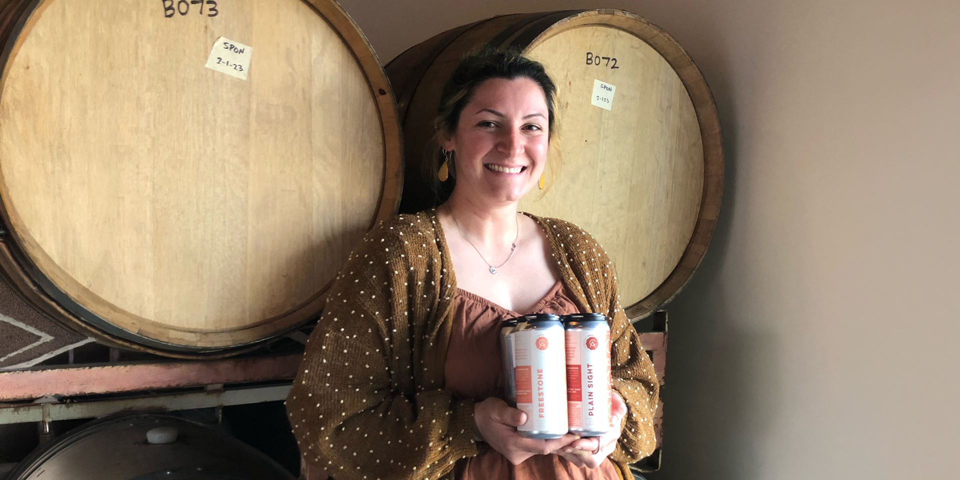 Bella Perez standing in front of two beer barrels, smiling, holding two cans of beer