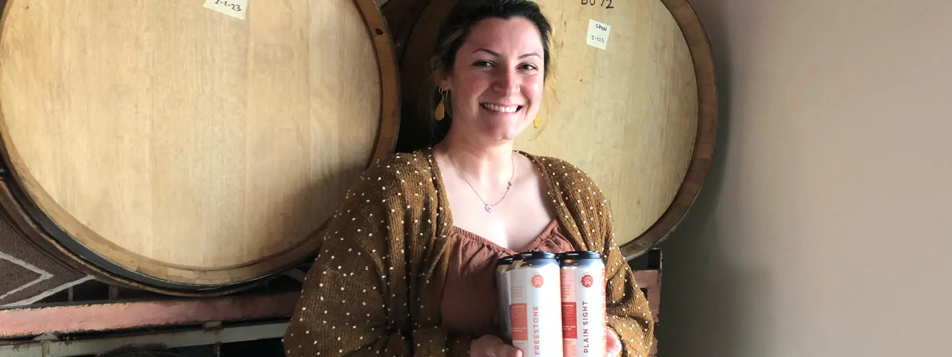 Bella Perez standing in front of two beer barrels, smiling, holding two cans of beer