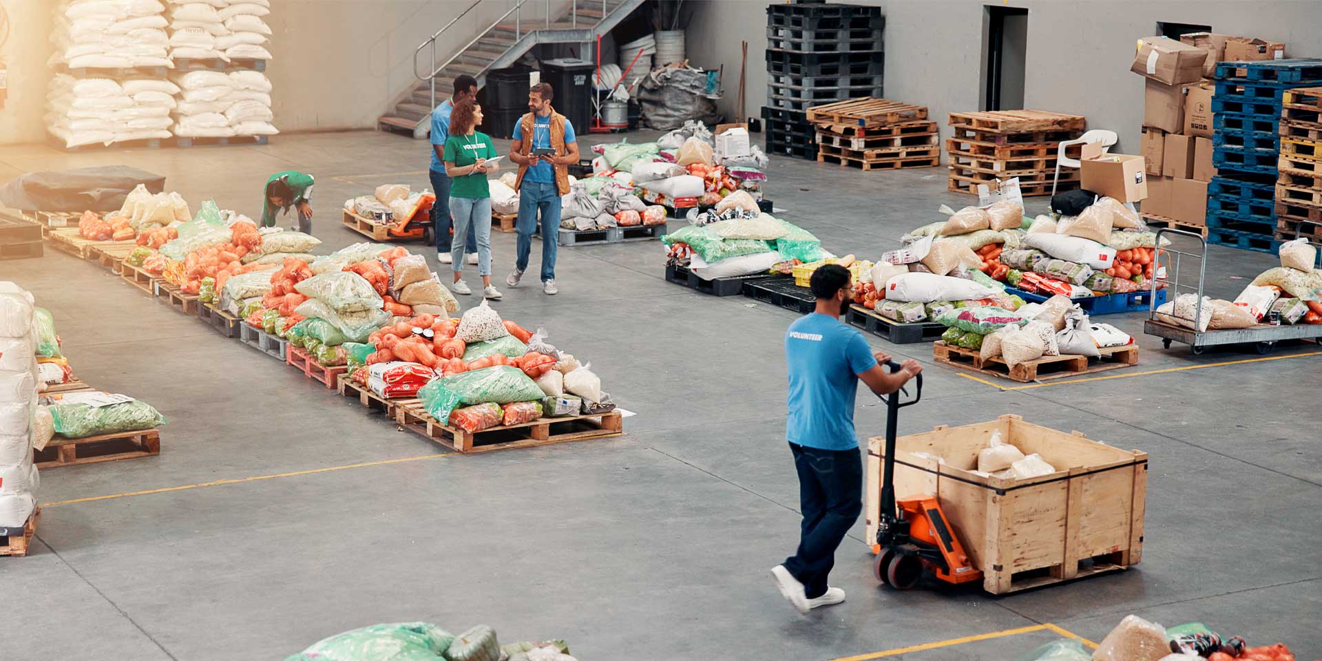 Volunteers organising and moving pallets of food supplies in a large warehouse with bags of vegetables and grains.
