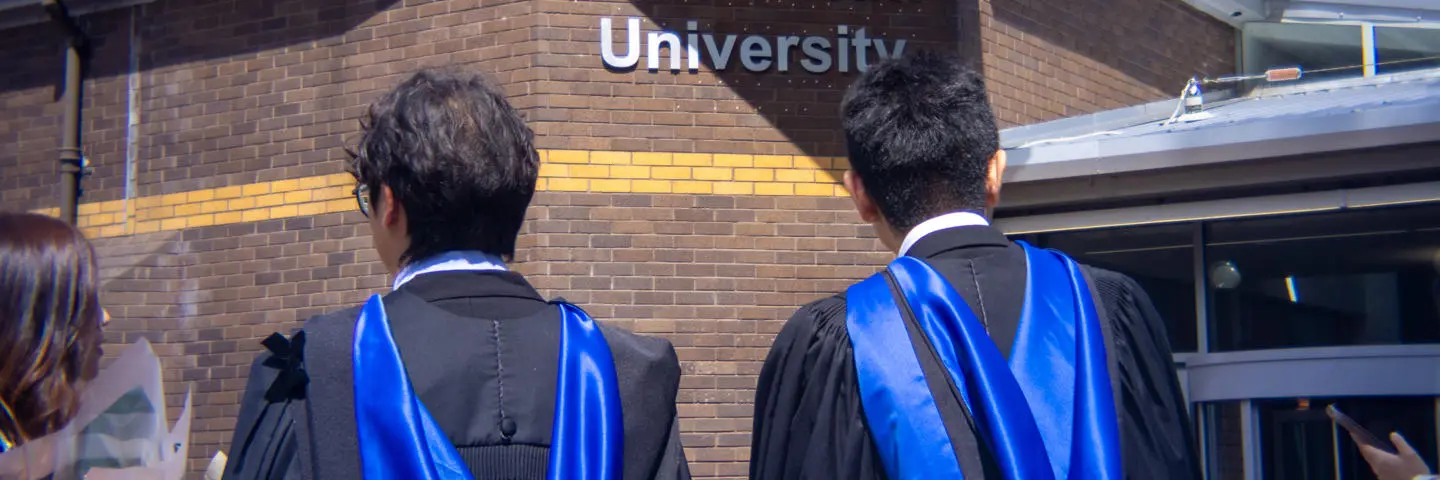 Shot shows two students standing in front of a sign that reads Heriot-Watt University.