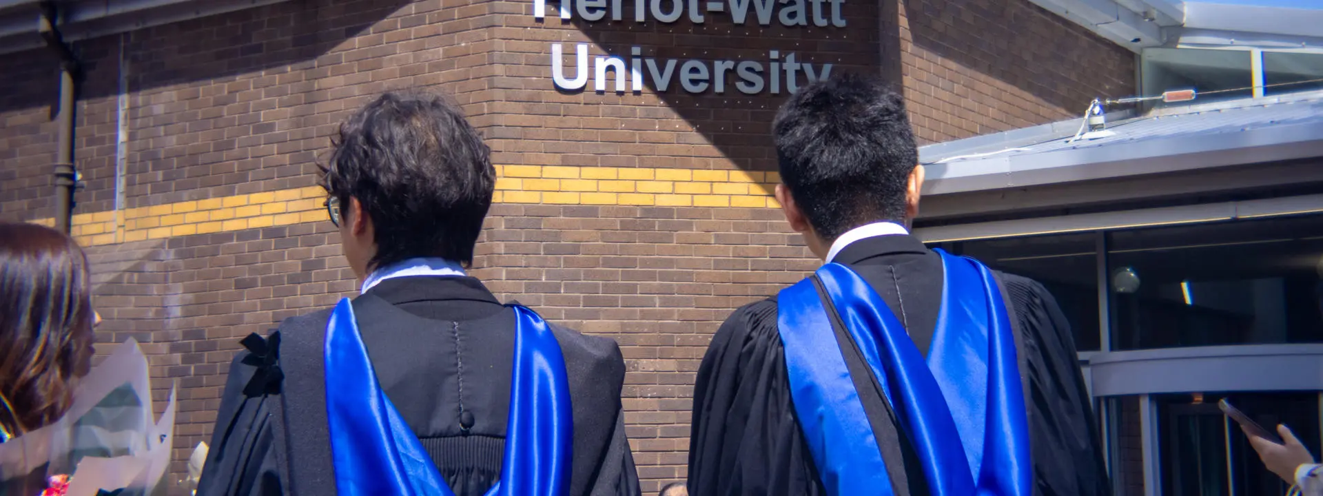 Shot shows two students standing in front of a sign that reads Heriot-Watt University.