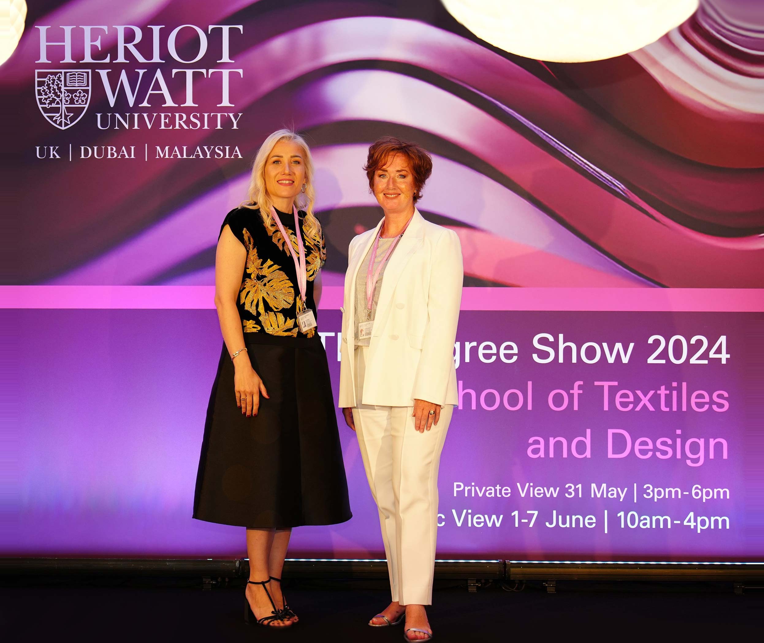 Two women standing in front of a banner that reads Herriot Watt School of Textiles and Design