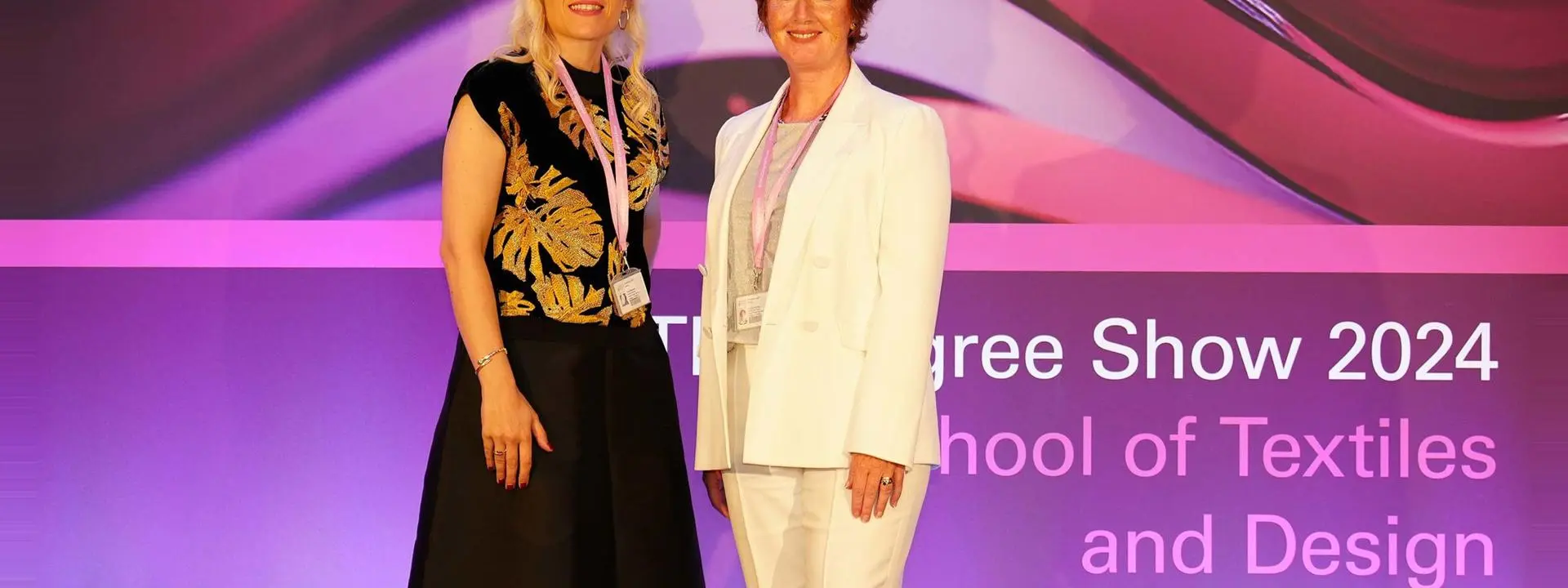 Two women standing in front of a banner that reads Herriot Watt School of Textiles and Design