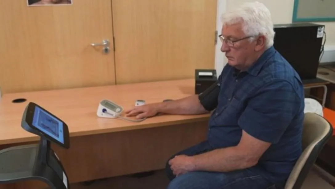 A man sits at a doctor's desk beside a Temi robot, with medical monitoring equipment