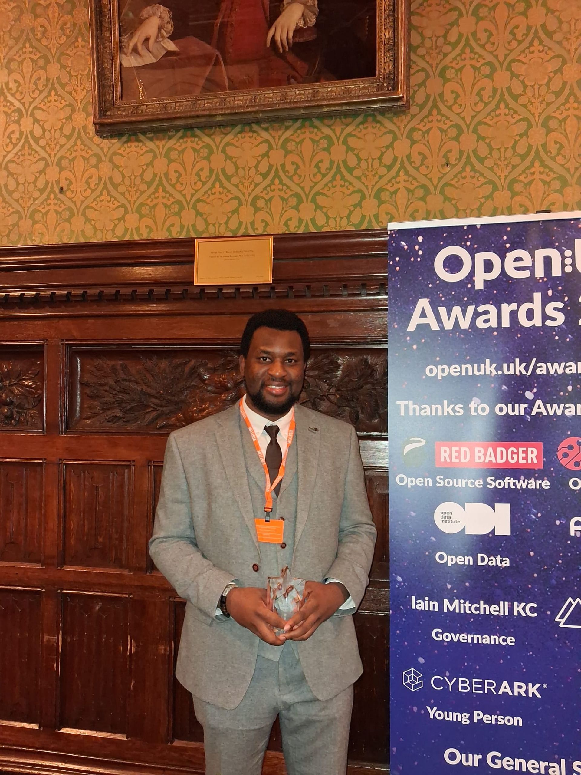 A person holding a glass award, wearing a suit, beside a vertical banner for the OpenUK Awards
