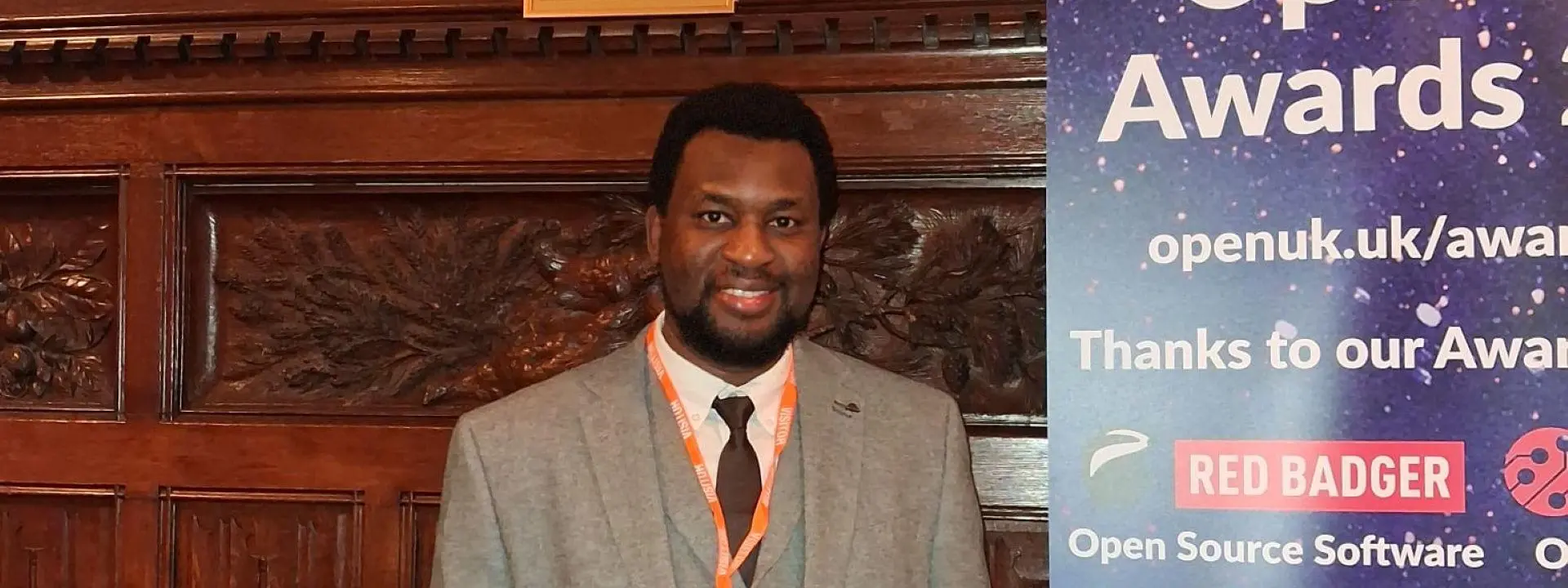 A person holding a glass award, wearing a suit, beside a vertical banner for the OpenUK Awards