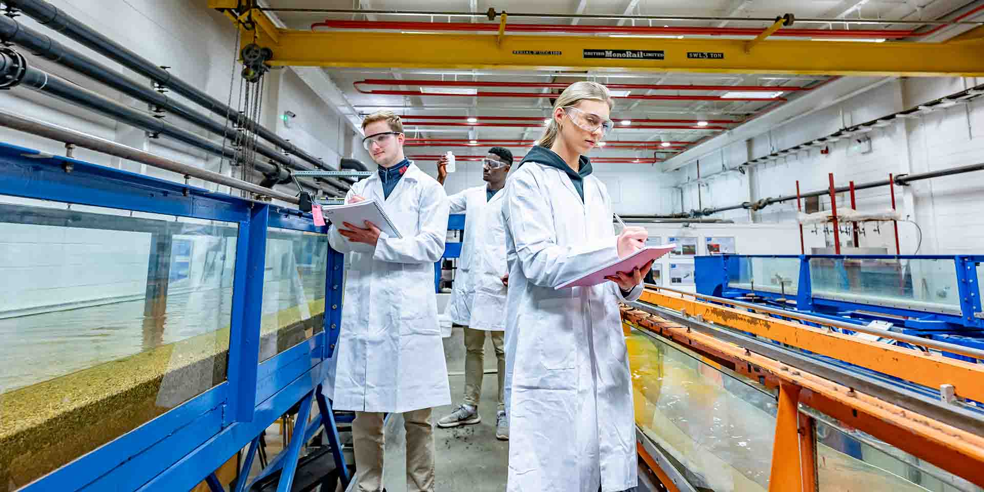 Image shows three students in lab coats, carrying out research. 