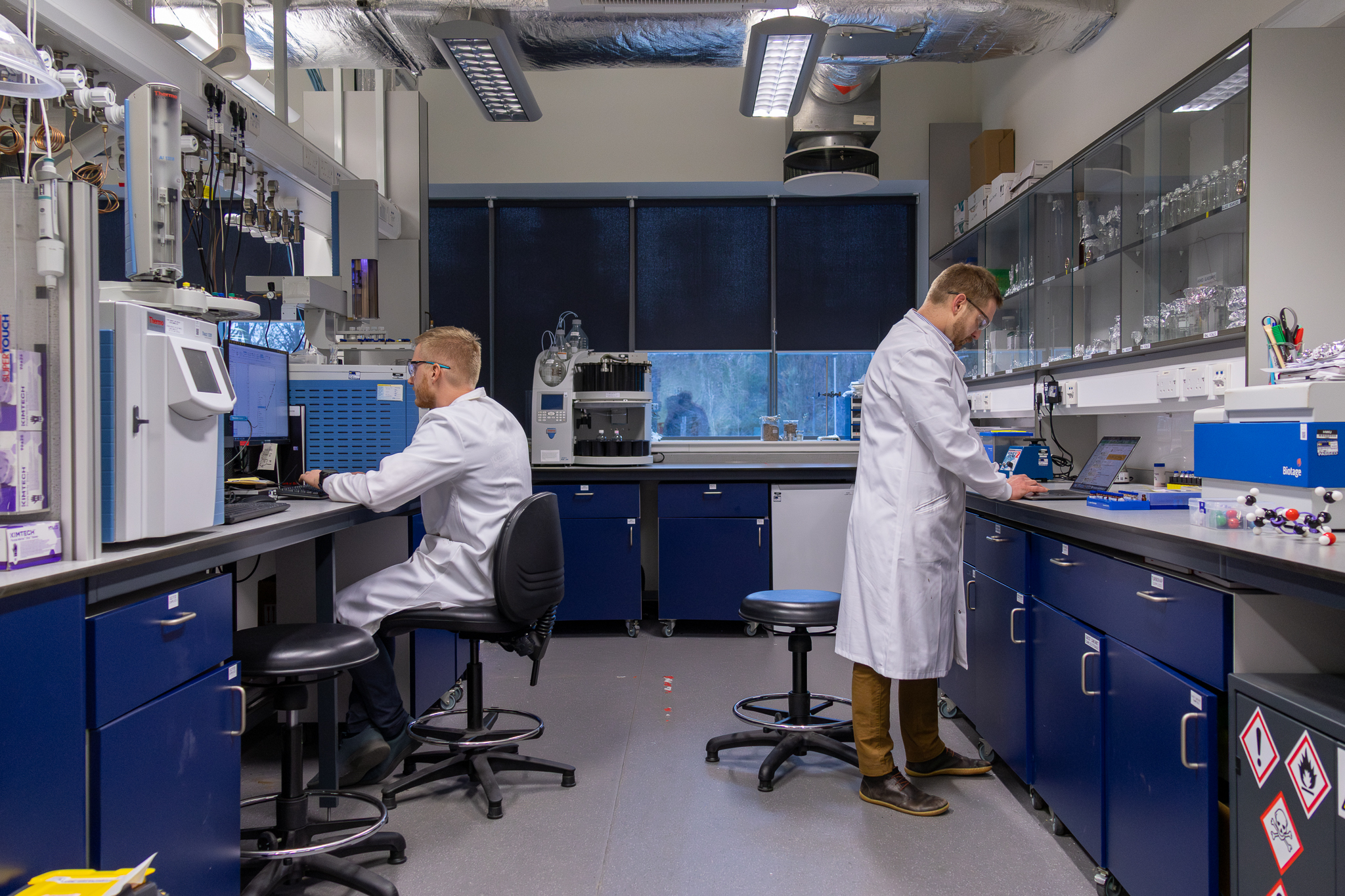 Two scientists in white coats stood opposite each other in a laboratory setting looking at computers and lab equipment. 