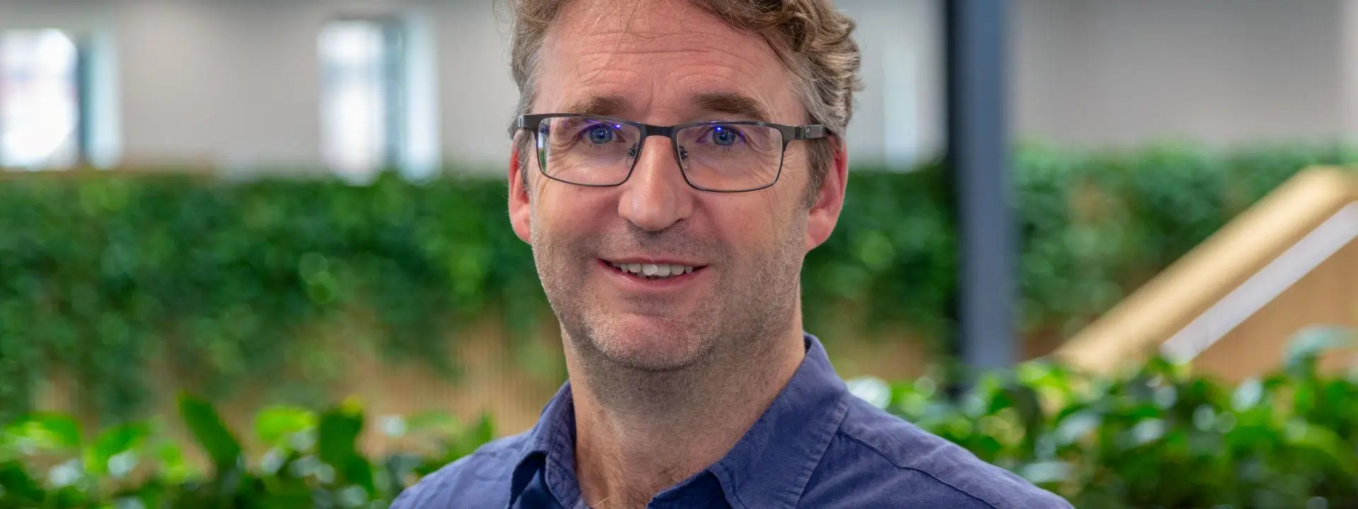 A man with wavy light brown hair, wearing glasses and a blue collared shirt, smiles warmly.