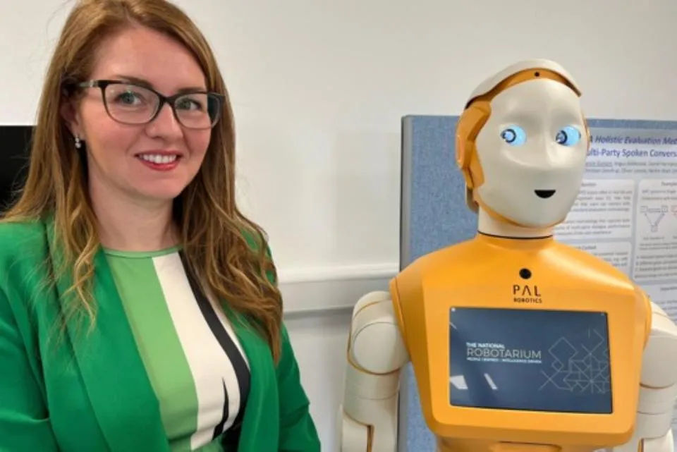 A smiling woman wearing glasses and a green jacket stands beside an orange-and-white humanoid robot with a touchscreen on its chest.