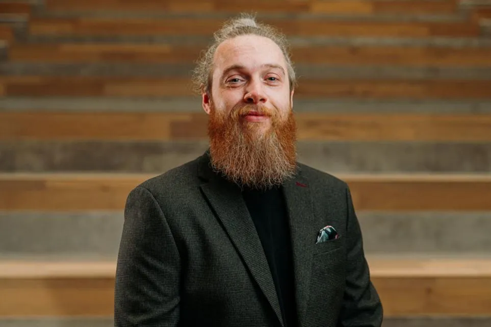 A man wearing a dark blazer and black high-neck top, standing in front of tiered wooden seating