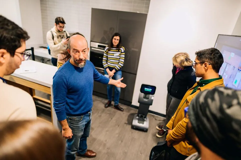 A group of people stand in a modern lab or office kitchen space. The setting is a technical research discussion in robotics