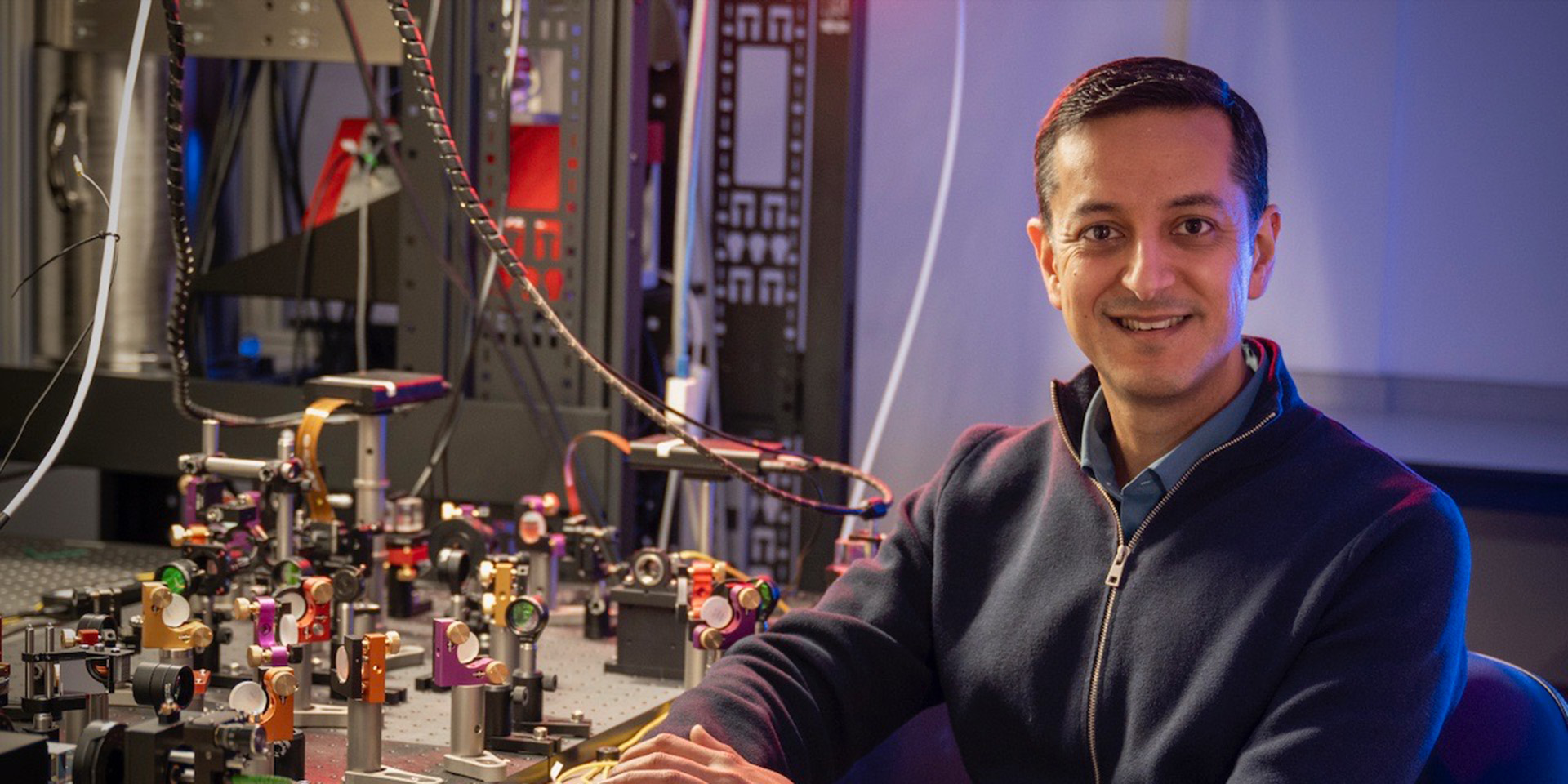 Professor Mehul Malik sitting beside laboratory bench with research apparatus 