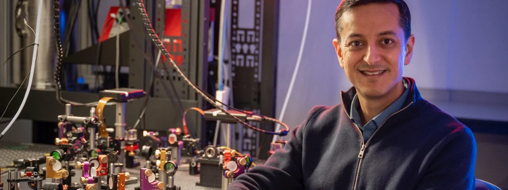 Professor Mehul Malik sitting beside laboratory bench with research apparatus