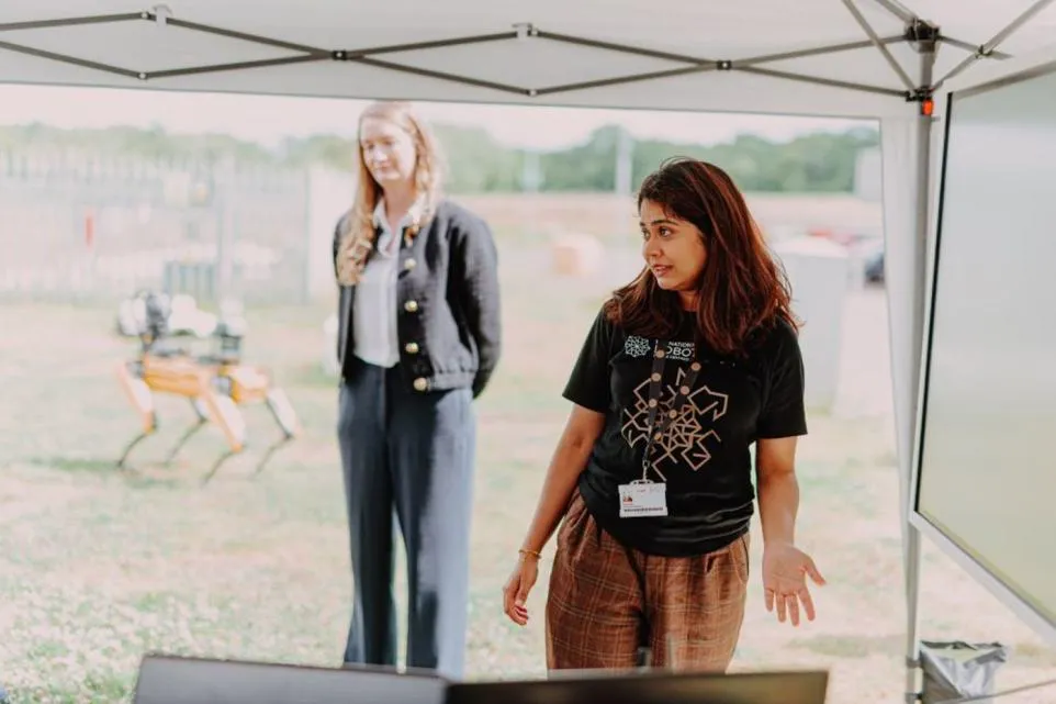 Two people standing under a gazebo; one wears a black National Robotarium t-shirt. In the background, a yellow robotic dog is visible along with outdoor scenery.