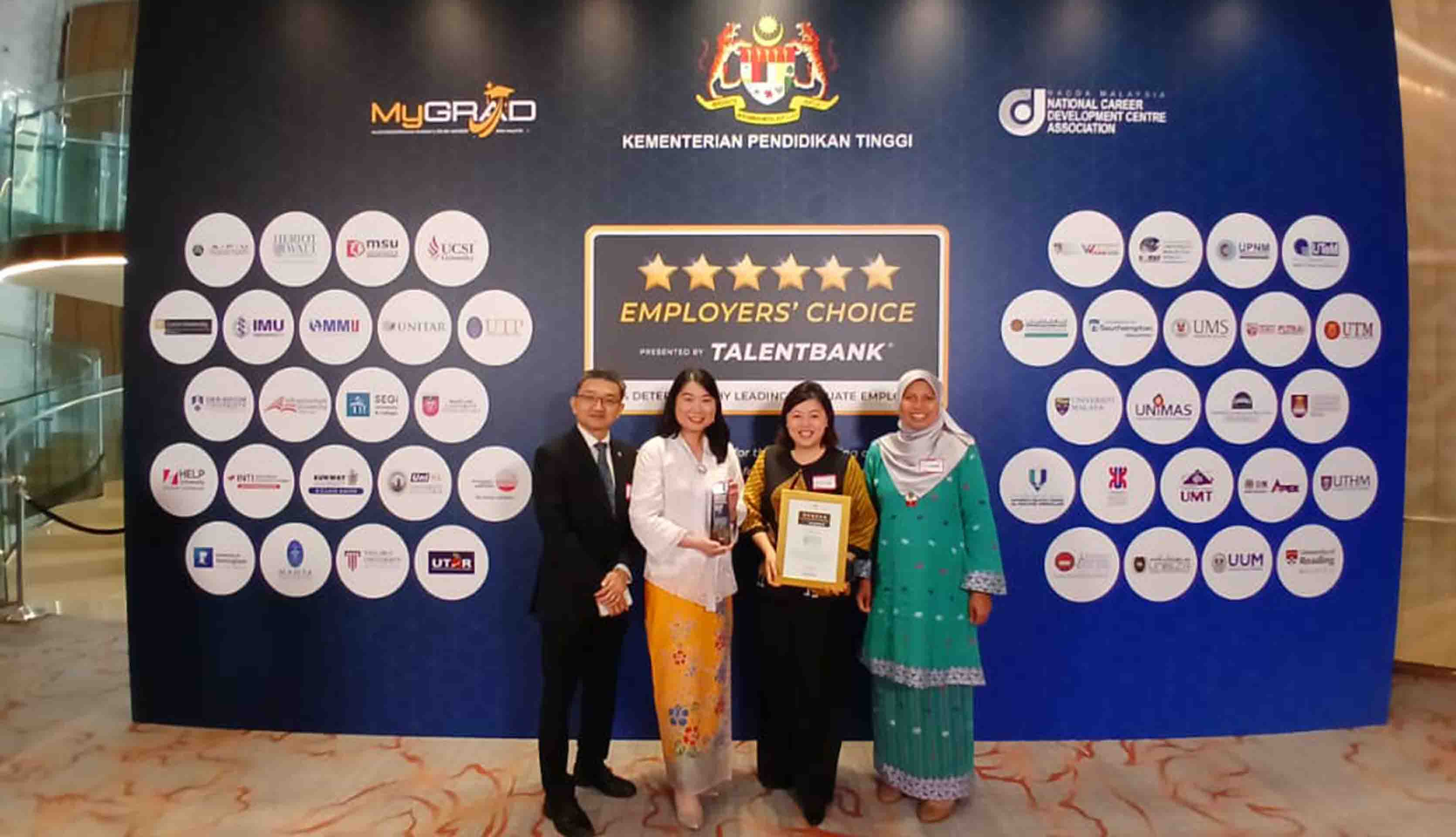 Image shows four people standing in front of a large banner with once holding a framed certificate. 