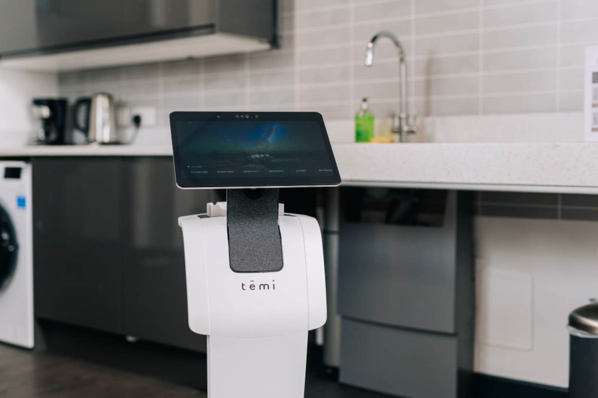 A white Temi service robot with a large touchscreen stands in the foreground of a modern kitchen