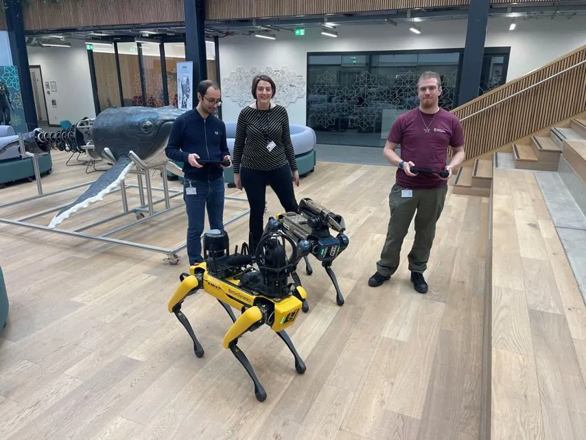 A group of three people stands in a modern indoor space with wooden flooring, open staircases, and contemporary furniture. Two of them are holding controllers while interacting with a yellow Boston Dynamics Spot robot, equipped with additional sensors and cameras.