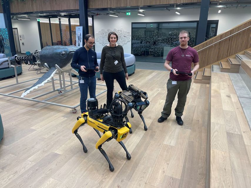 Research technicians from UKRAS-STEPs on secondment at the National Robotarium, alongside Project Manager Catherine Ciarka-White A group of three people stands in a modern indoor space with wooden flooring, open staircases, and contemporary furniture. Two of them are holding controllers while interacting with a yellow Boston Dynamics Spot robot, equipped with additional sensors and cameras.