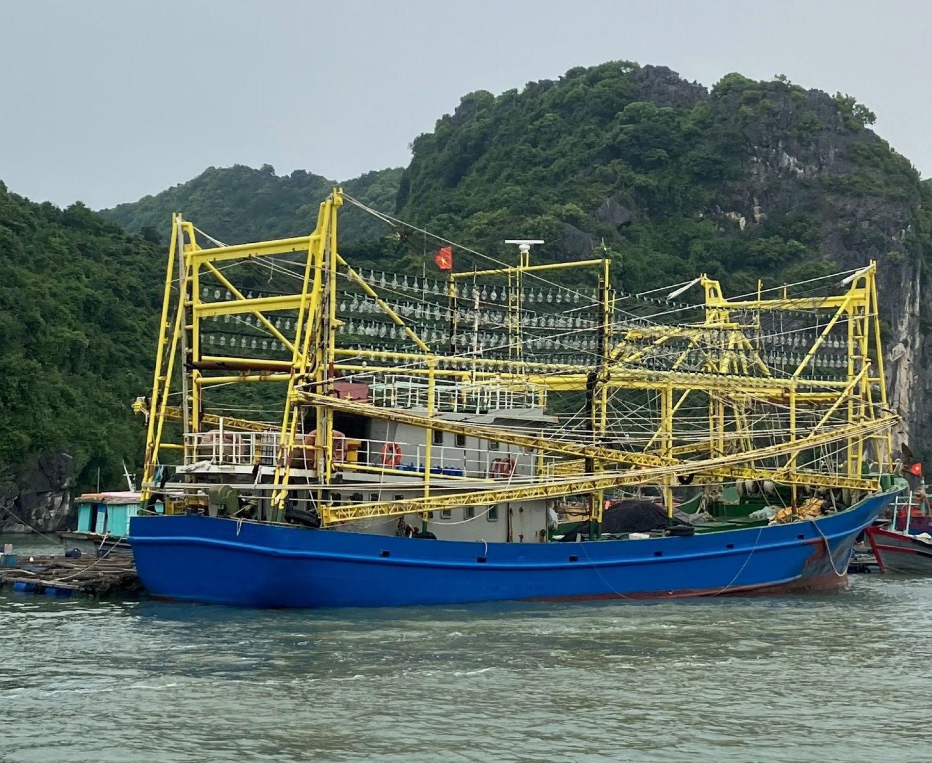 A large blue fishing boat is moored on calm water in front of forested limestone hill