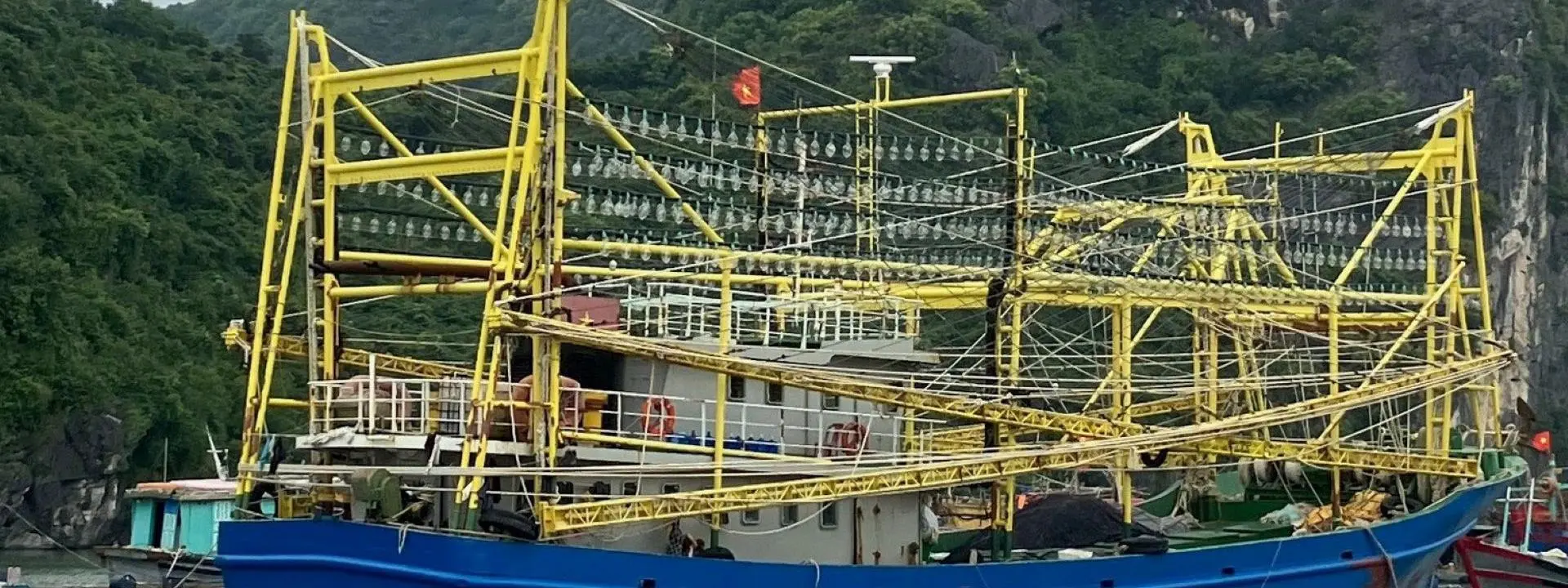 A large blue fishing boat is moored on calm water in front of forested limestone hill