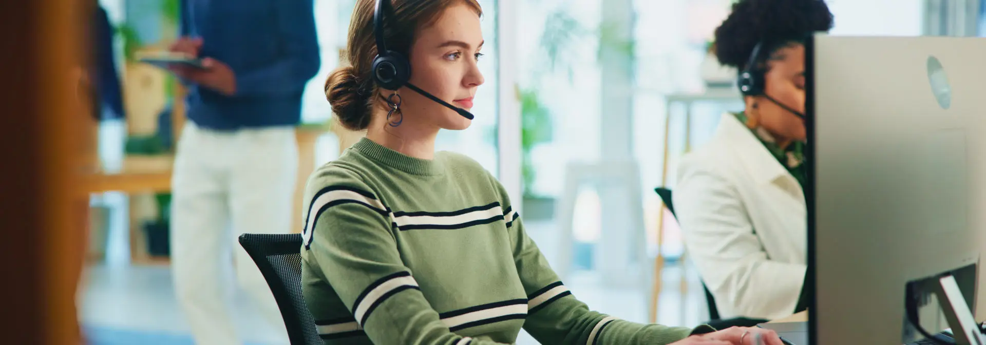 A woman wearing a headset sits at a computer on the phone.