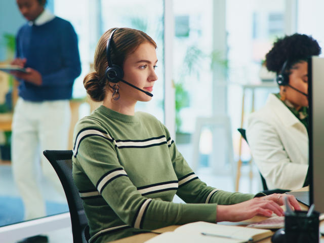 A woman wearing a headset sits at a computer on the phone.