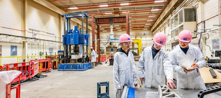 Three individuals wearing lab coats and safety helmets in warehouse.