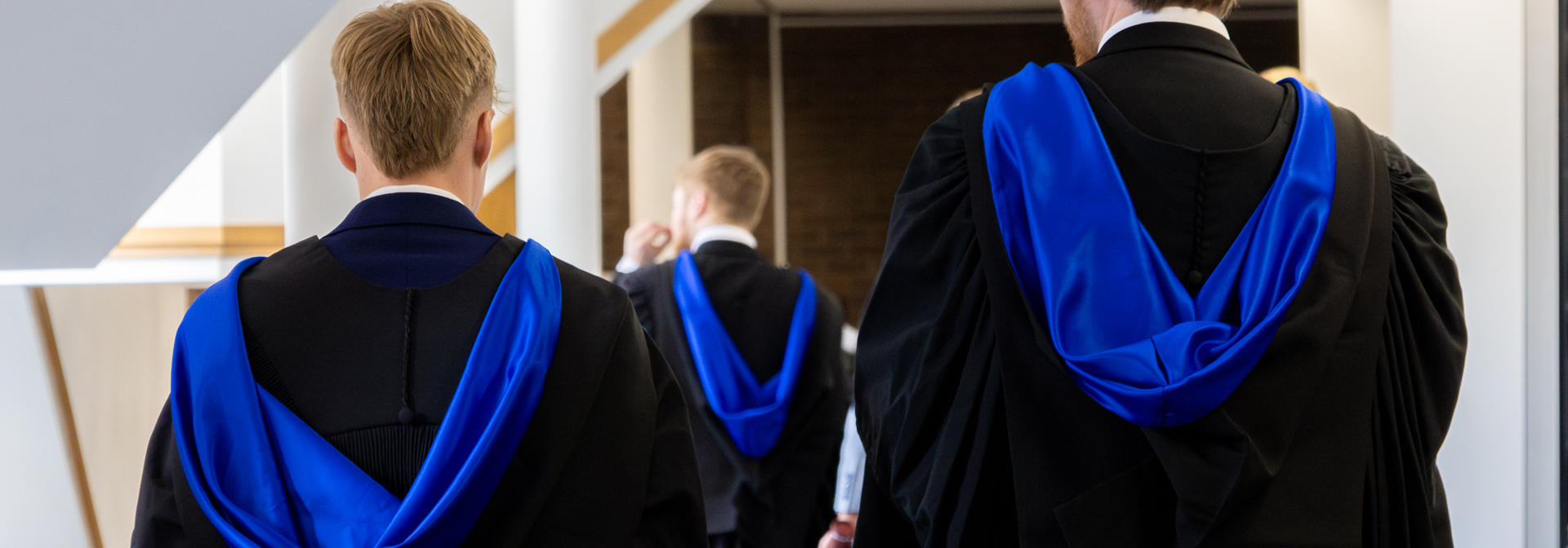 Three individuals wearing graduation robes walking down a corridor.