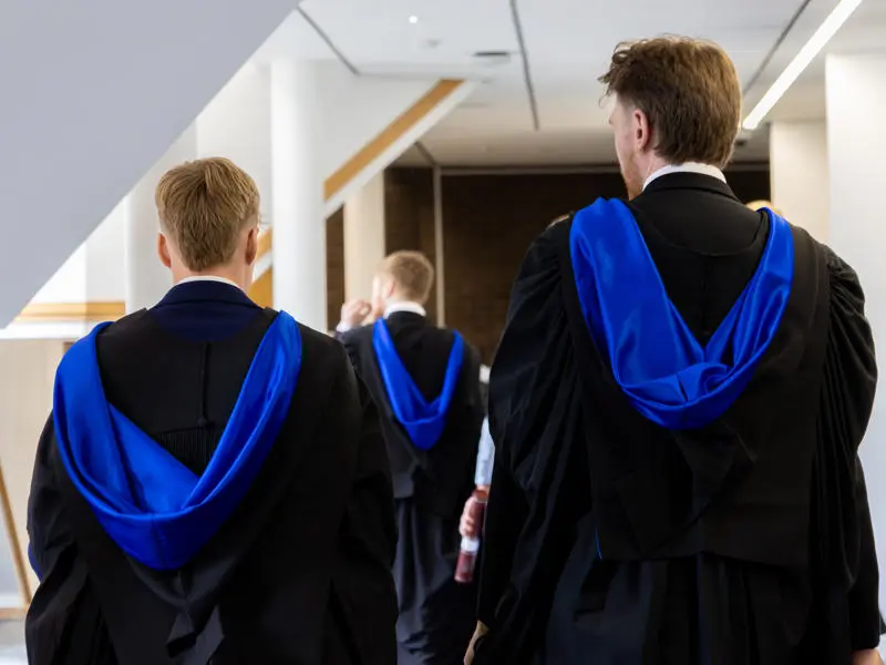 Three individuals wearing graduation robes walking down a corridor.