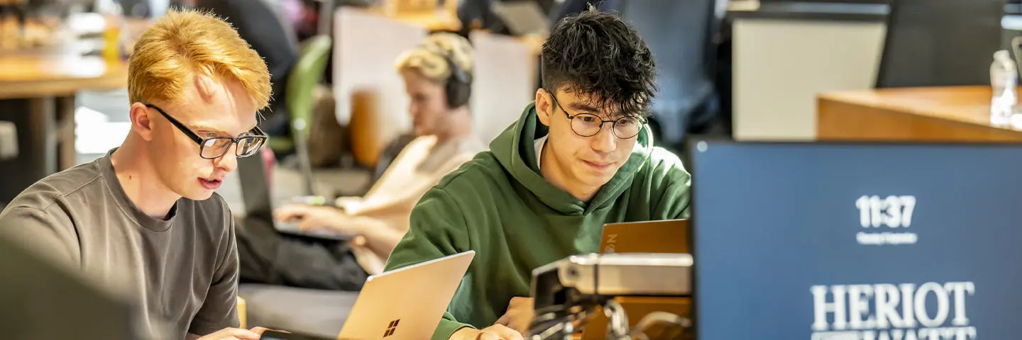 Two focused students study together using laptops in a relaxed, modern indoor setting.