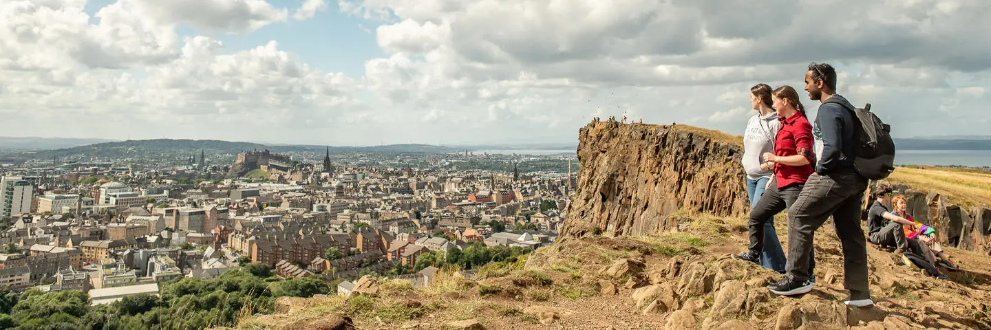 Three individuals stand atop a hill and look over at a city below.