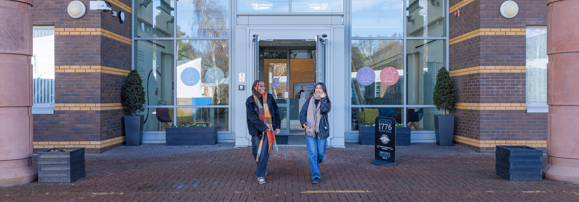Two students outside the EBS building, with large glass doors and potted plants flanking the entrance.