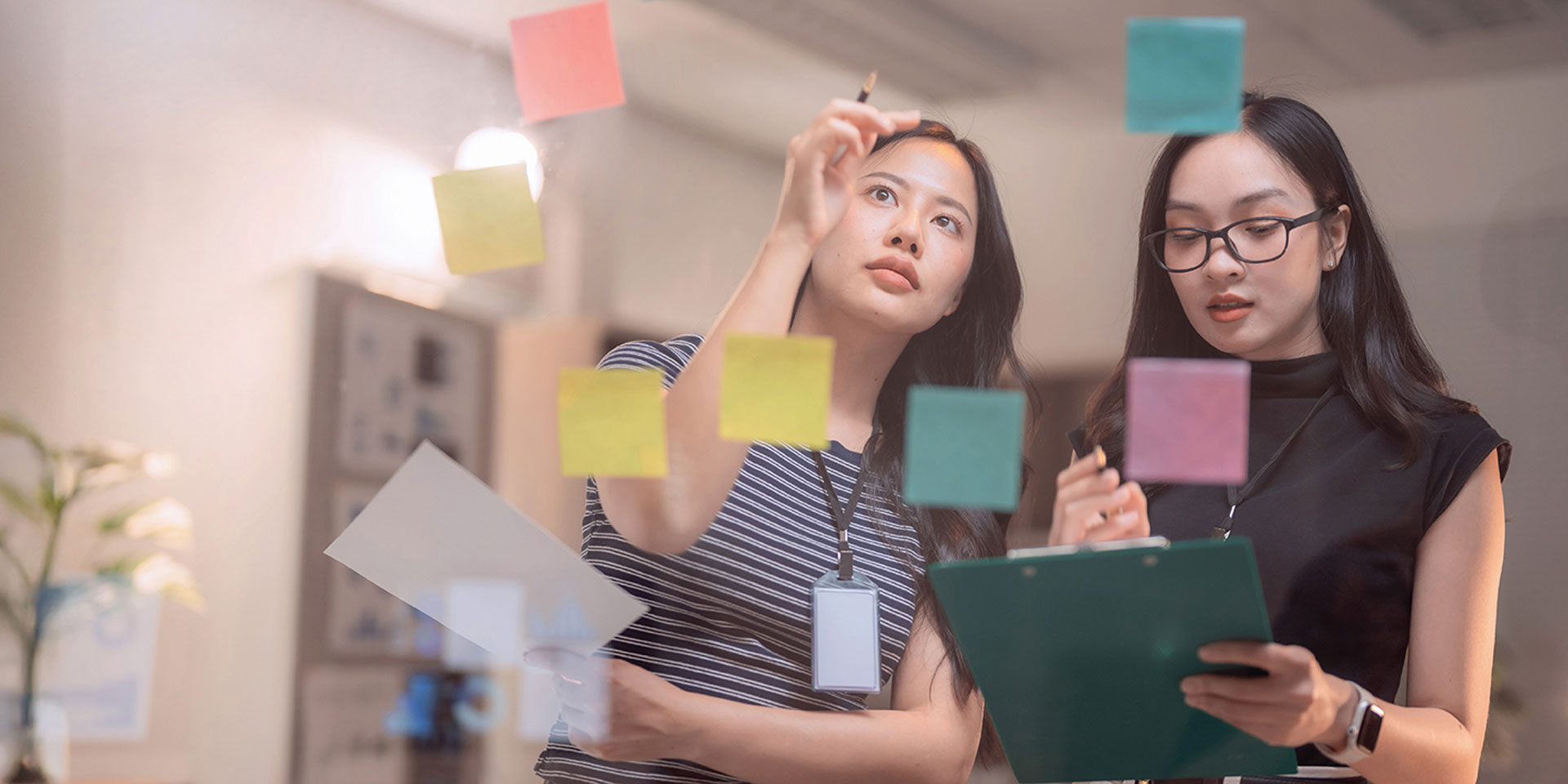 Two women examine colorful sticky notes arranged on a wall, discussing ideas and collaborating on a project.